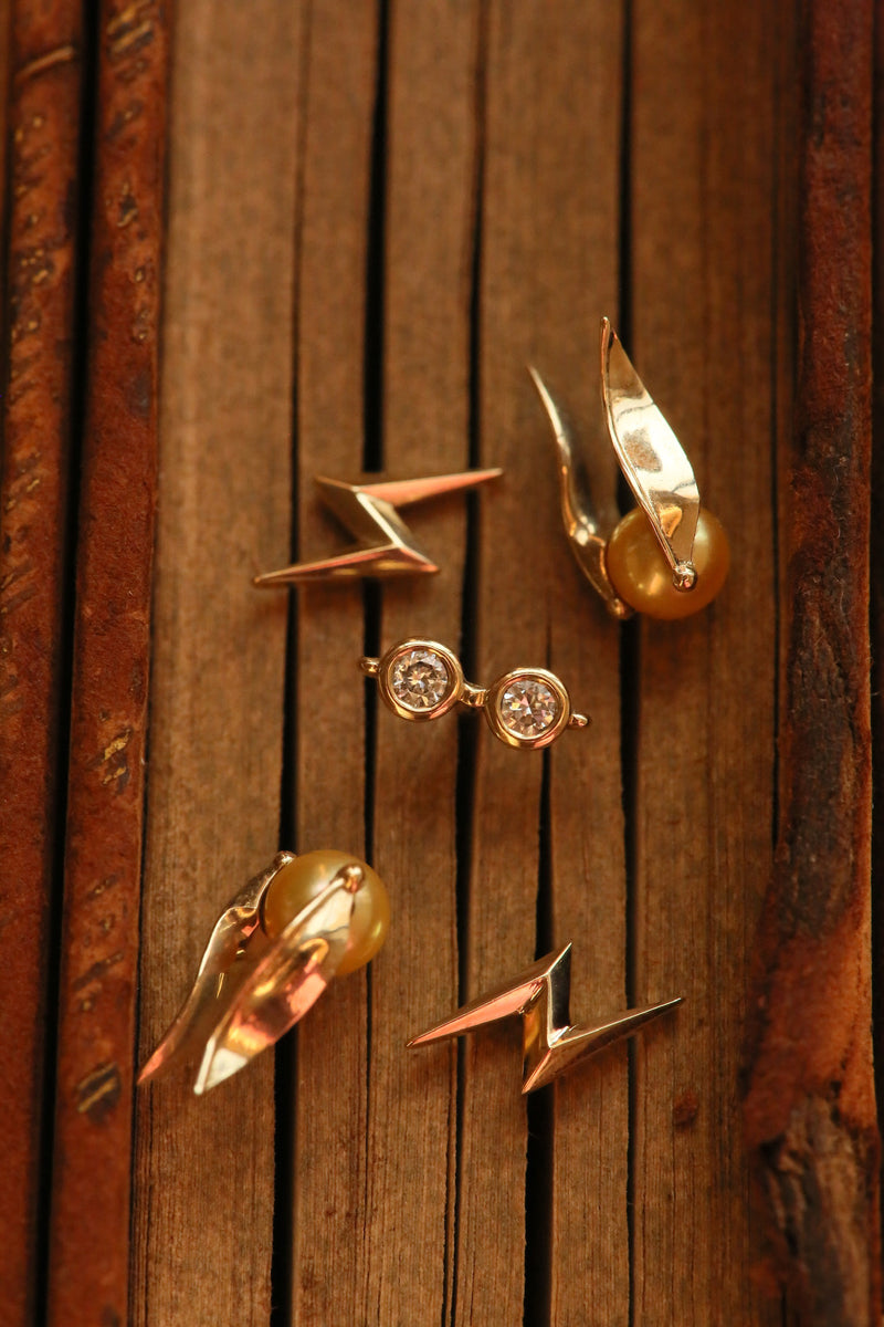 Officially Licensed Harry Potter fine jewelry by Freeman Design in solid 14k gold sitting on a rustic wooden background. Jewelry featured: Harry Potter Glasses Earrings, Lightning Bolt Stud Earrings, and Golden Pearl Snitch Earrings. Also available in various other metals. 