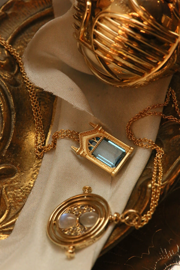 Golden Snitch Ring Box, Mirror of Erised necklace, and Time Turner necklace cast in 14k yellow gold nestle on a piece of buff fabric in a golden dish. All of these officially licensed Harry Potter fine jewelry pieces are available in other precious metals.