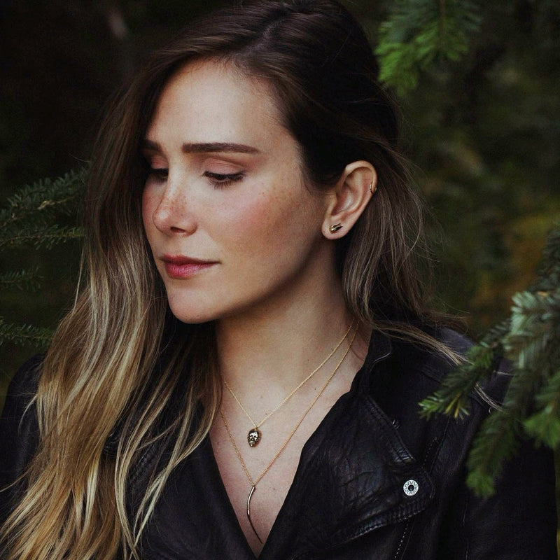 Portrait of a woman wearing officially licensed Harry Potter jewelry in front of pine boughs. Jewelry featured: Golden Pearl Snitch Earrings, Bellatrix LeStrange death eater mask, and Bellatrix LeStrange Wand in solid 14k yellow gold. Also available in other metals.