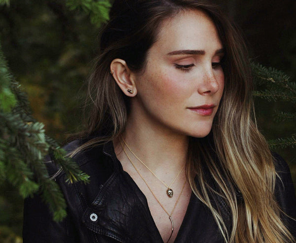 Portrait of a woman wearing officially licensed Harry Potter jewelry in front of pine boughs. Jewelry featured: Golden Pearl Snitch Earrings, Bellatrix LeStrange death eater mask, and Bellatrix LeStrange Wand, all in solid 14k yellow gold. Also available in other metals.